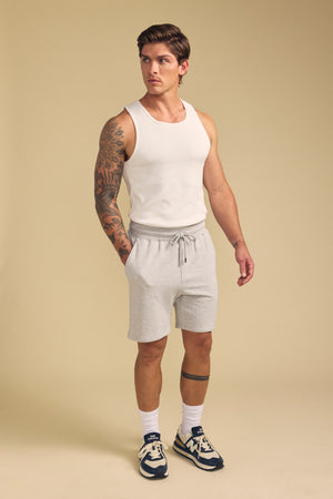 A man in a white tank top, Velvet by Graham & Spencer QUENTIN SWEATSHORT in light gray, white socks, and navy sneakers stands against a beige background.
