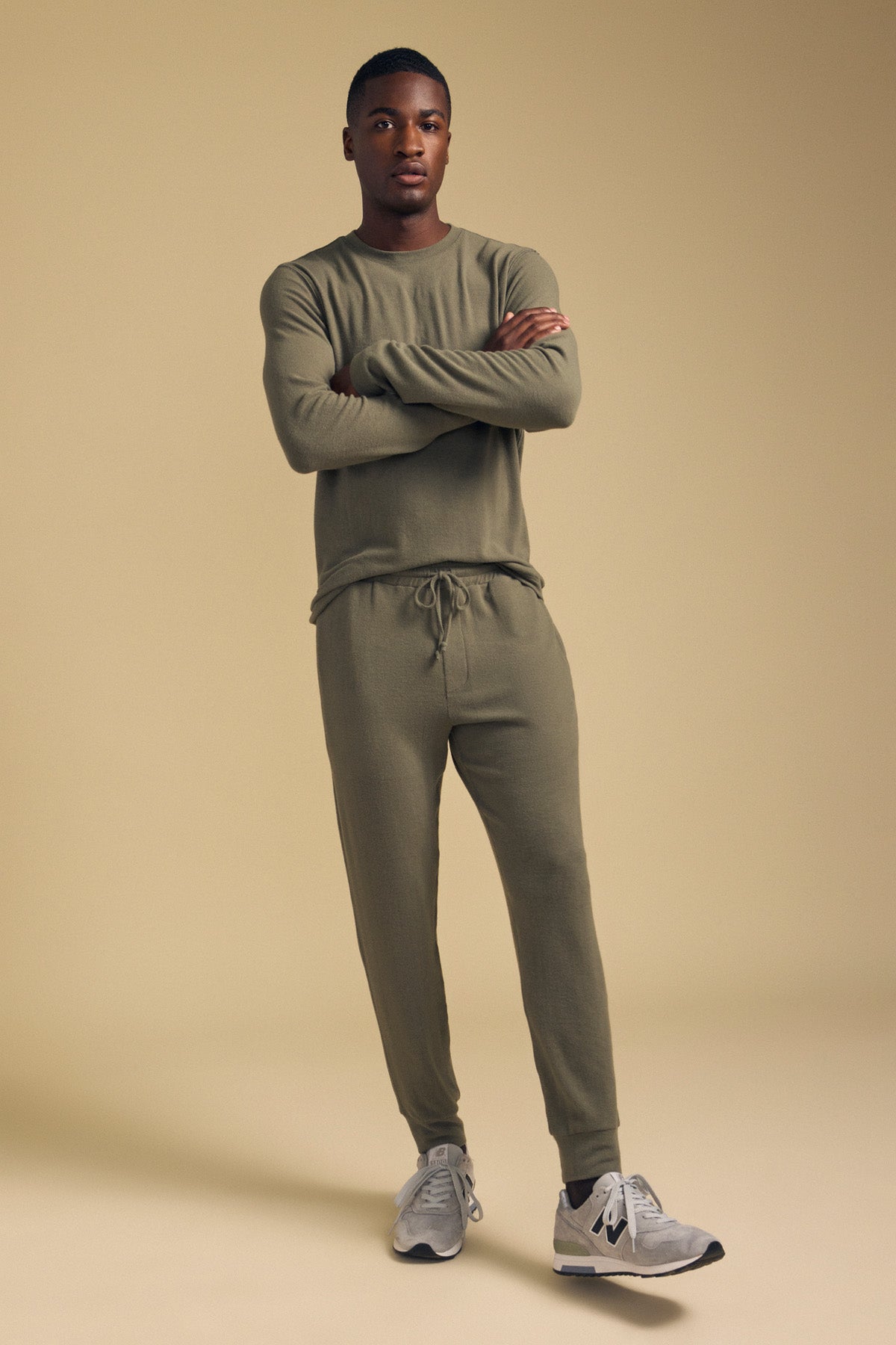 A man stands with arms crossed, wearing the Velvet by Graham & Spencer REDDEN CREW PULLOVER in olive green and joggers, paired with gray sneakers, against a beige background—perfect for everyday wear.-42092504350913
