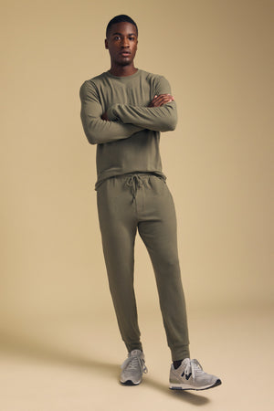 A man stands with arms crossed, wearing the Velvet by Graham & Spencer REDDEN CREW PULLOVER in olive green and joggers, paired with gray sneakers, against a beige background—perfect for everyday wear.