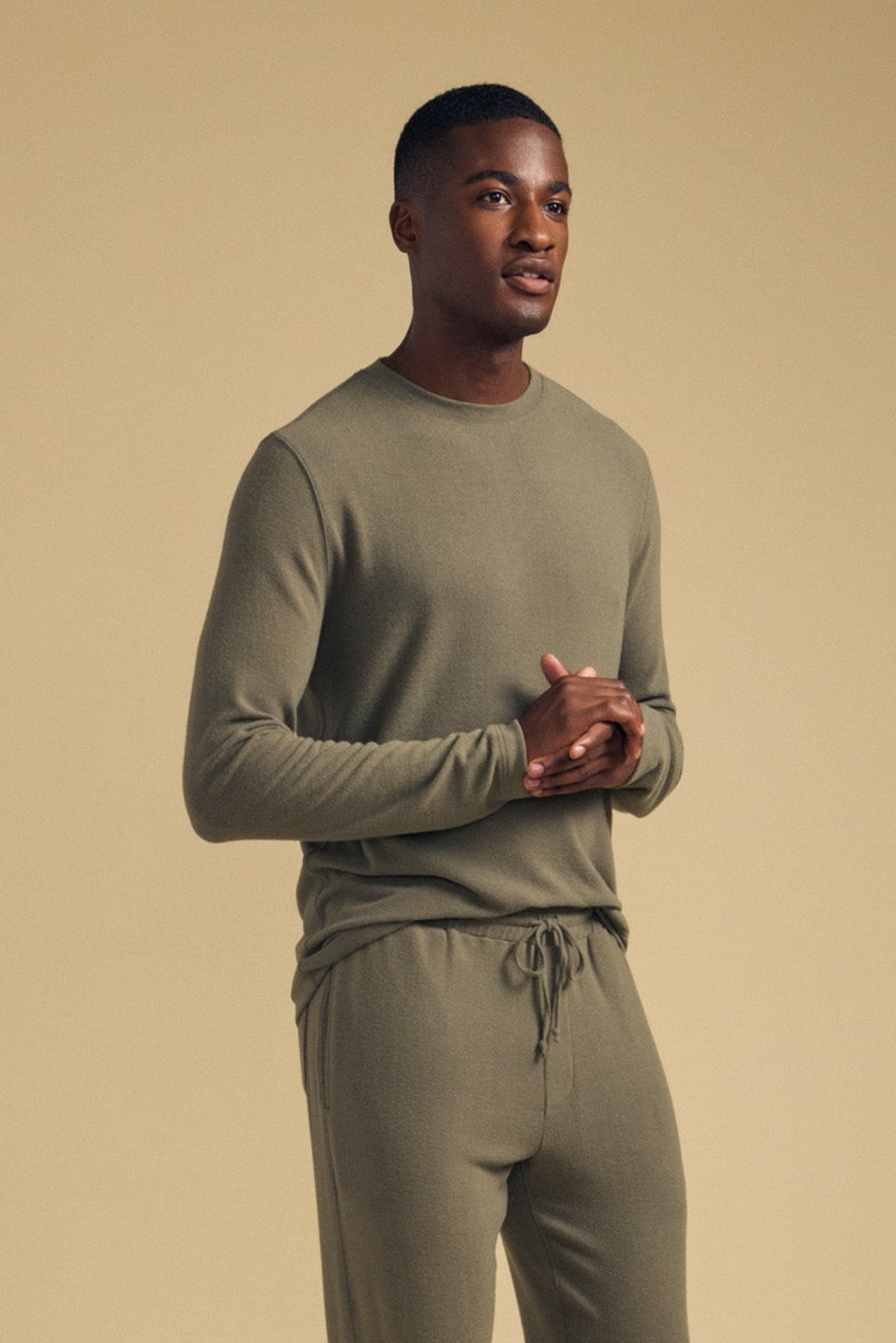 A man stands against a beige background wearing the REDDEN CREW PULLOVER by Velvet by Graham & Spencer—an olive green long-sleeve shirt crafted from cozy jersey fabric—paired with matching jogger pants and gray sneakers for an ideal everyday look.