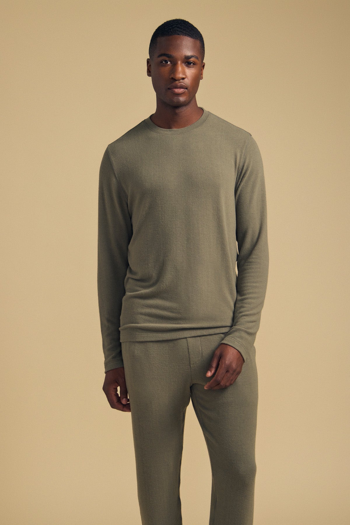 A man stands against a plain beige background, wearing the Velvet by Graham & Spencer REDDEN CREW PULLOVER in olive green paired with matching pants, looking directly at the camera.