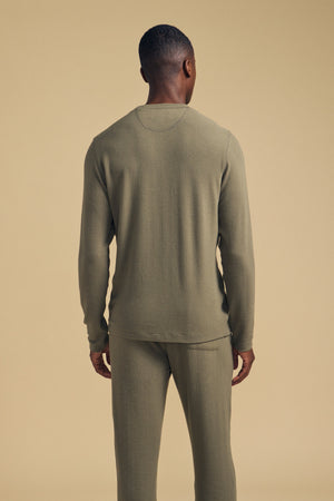 A man stands facing away from the camera, wearing the Velvet by Graham & Spencer REDDEN CREW PULLOVER in olive green with matching pants against a beige background—perfect for everyday wear.
