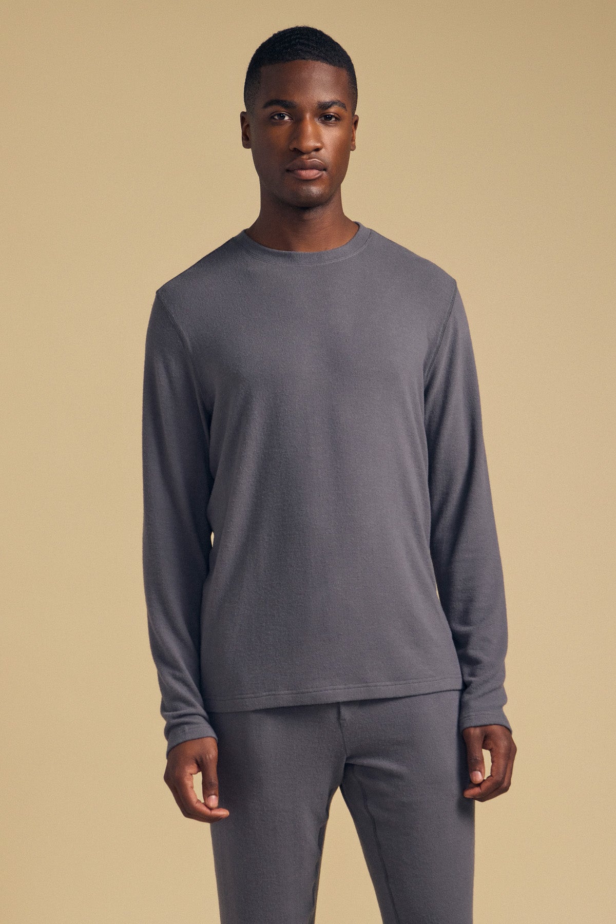 A person stands facing the camera, wearing the Velvet by Graham & Spencer REDDEN CREW PULLOVER—a matching long-sleeve gray shirt and gray pants—against a plain beige background.-41859788439745