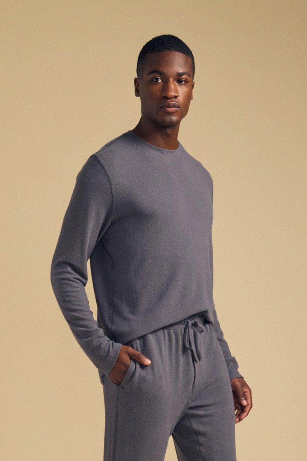 A man stands against a beige background, wearing the Velvet by Graham & Spencer REDDEN CREW PULLOVER and matching grey drawstring pants, with one hand in his pocket.