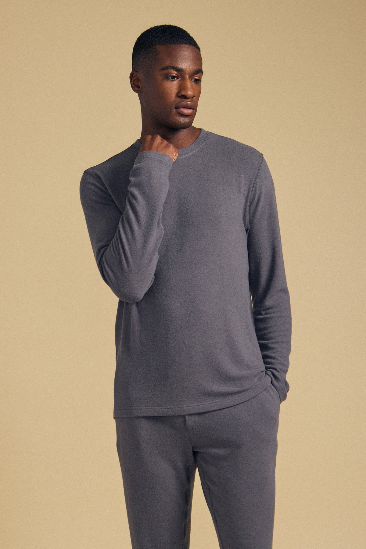 A man wearing the REDDEN CREW PULLOVER by Velvet by Graham & Spencer, paired with matching grey pants, stands against a plain beige background with one hand near his chin.