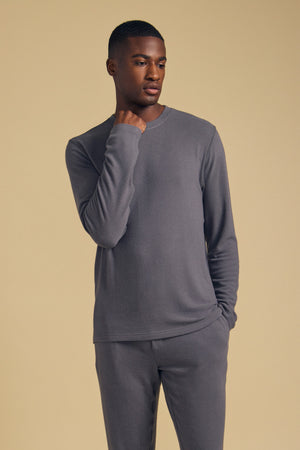 A man wearing the REDDEN CREW PULLOVER by Velvet by Graham & Spencer, paired with matching grey pants, stands against a plain beige background with one hand near his chin.