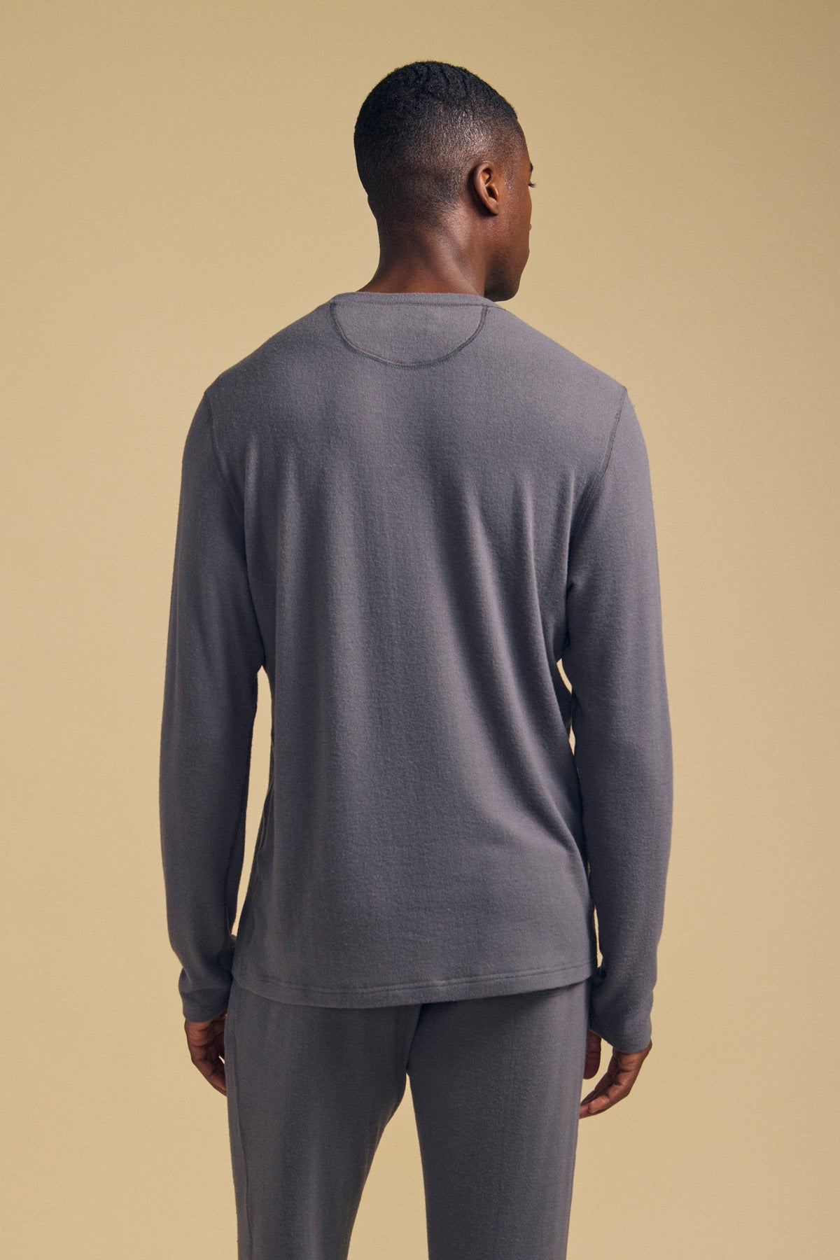 A person stands facing away from the camera, wearing the Velvet by Graham & Spencer REDDEN CREW PULLOVER in gray with matching pants, set against a plain beige background.