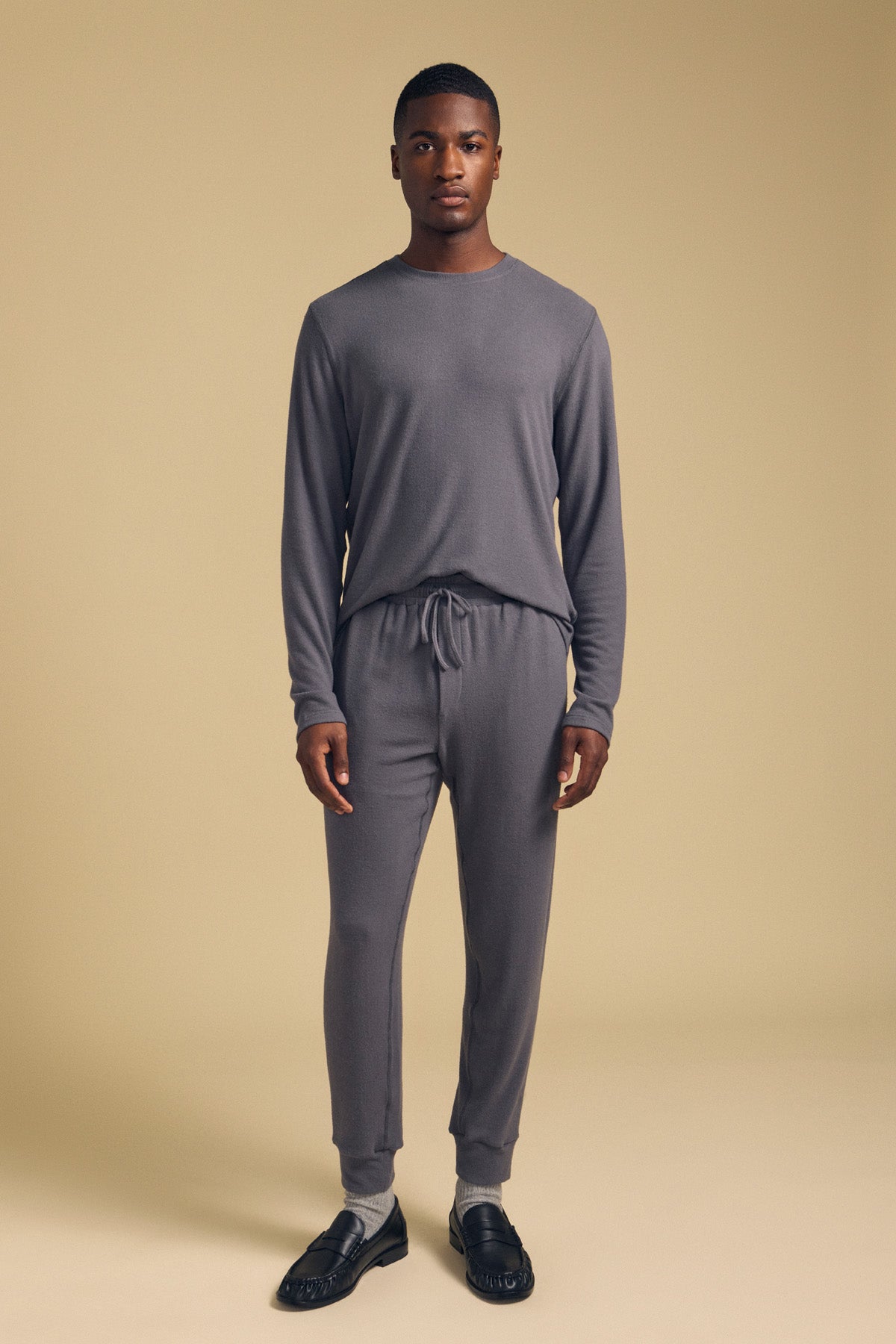 A man stands facing forward against a plain beige background, wearing the Velvet by Graham & Spencer REDDEN CREW PULLOVER and matching gray jogger pants, paired with black shoes and light-colored socks.