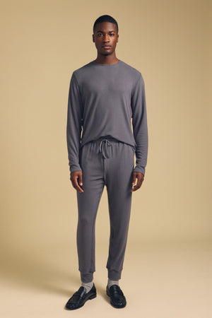 A man stands facing forward against a plain beige background, wearing the Velvet by Graham & Spencer REDDEN CREW PULLOVER and matching gray jogger pants, paired with black shoes and light-colored socks.