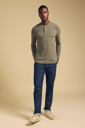 A man stands against a beige background with his hands in his pockets, wearing the TYLER HENLEY by Velvet by Graham & Spencer—an olive long-sleeve henley shirt crafted from soft jersey—paired with blue jeans and beige sneakers.
