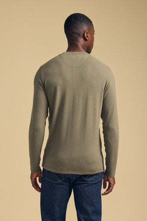 A person stands facing away from the camera, wearing the Velvet by Graham & Spencer TYLER HENLEY in olive green and dark blue jeans against a plain beige background.