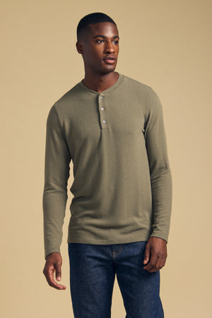A man stands against a beige background wearing the TYLER HENLEY long-sleeve olive-green jersey shirt by Velvet by Graham & Spencer and blue jeans.