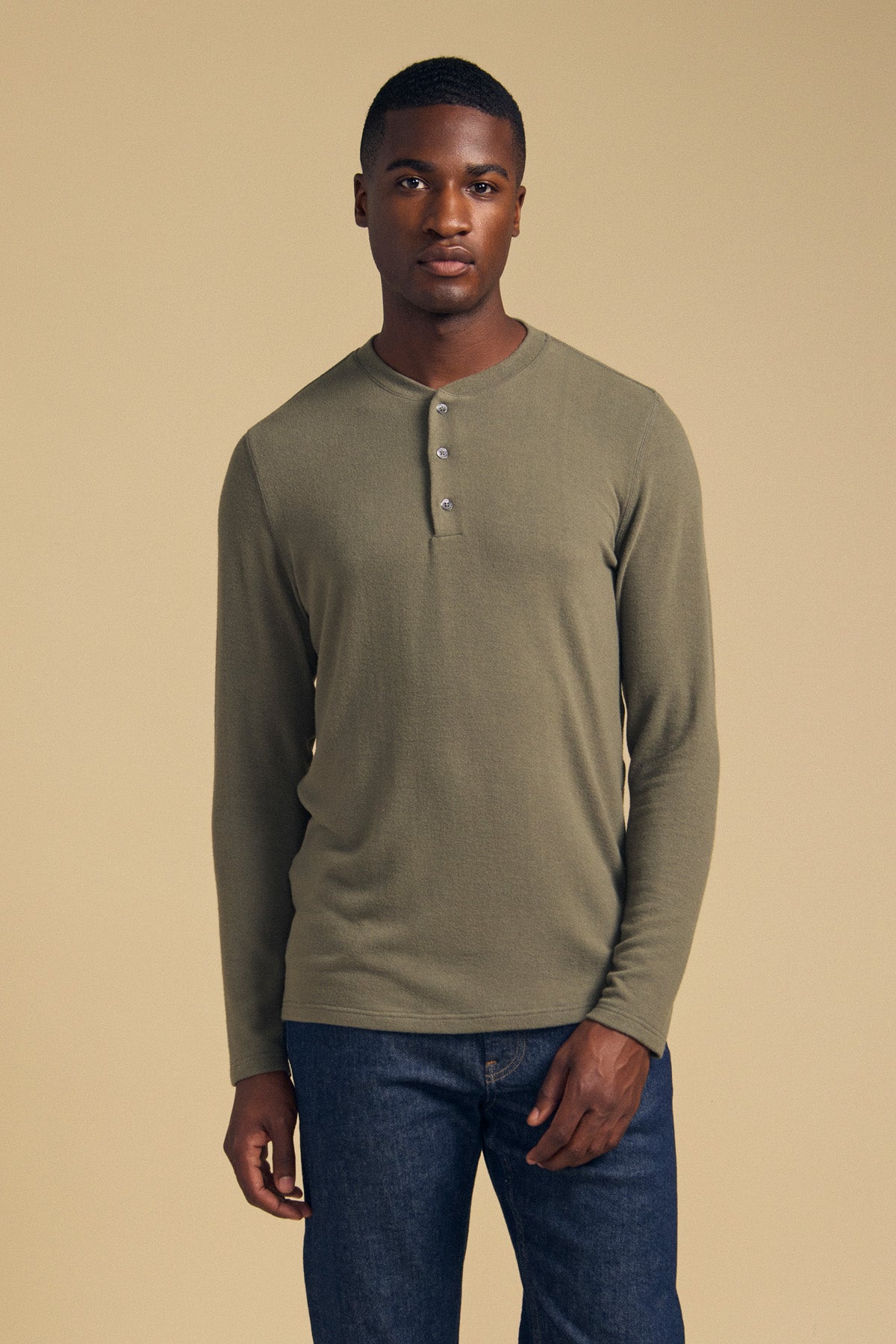 A man stands against a beige background, facing the camera with a neutral expression, wearing the TYLER HENLEY long sleeve jersey shirt in olive green by Velvet by Graham & Spencer and blue jeans.