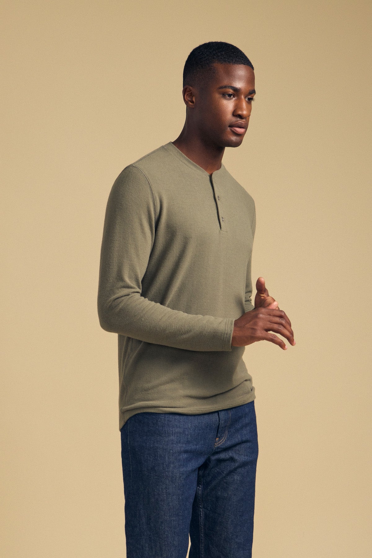 A man stands against a beige background, wearing the TYLER HENLEY long-sleeve shirt in olive green by Velvet by Graham & Spencer, paired with blue jeans.
