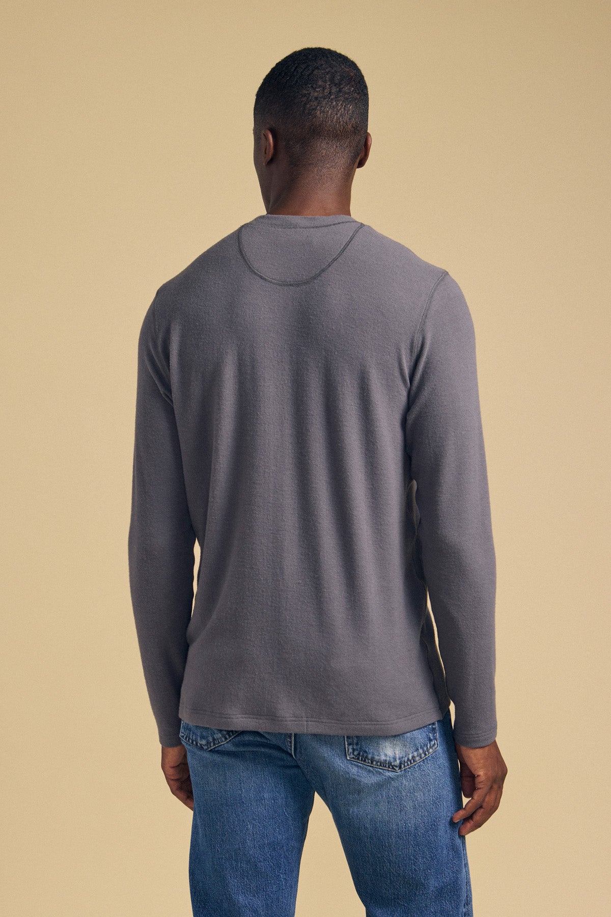 A person stands facing away against a beige background, wearing the Velvet by Graham & Spencer TYLER HENLEY in gray with blue jeans.