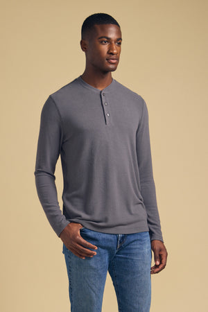 A person stands against a beige background wearing the Velvet by Graham & Spencer TYLER HENLEY in gray and blue jeans, looking slightly to the side with one hand in their pocket.