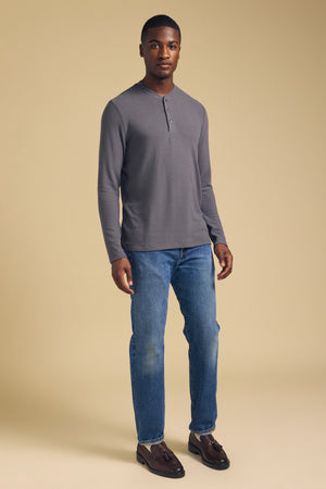 A man stands against a beige background wearing the TYLER HENLEY long sleeve jersey shirt by Velvet by Graham & Spencer, paired with blue jeans and brown shoes.