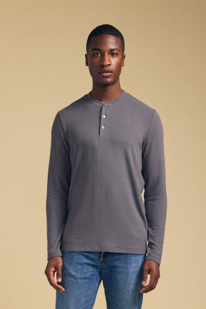 A man stands facing forward, wearing the TYLER HENLEY long-sleeve jersey shirt by Velvet by Graham & Spencer in gray and blue jeans against a plain beige background.