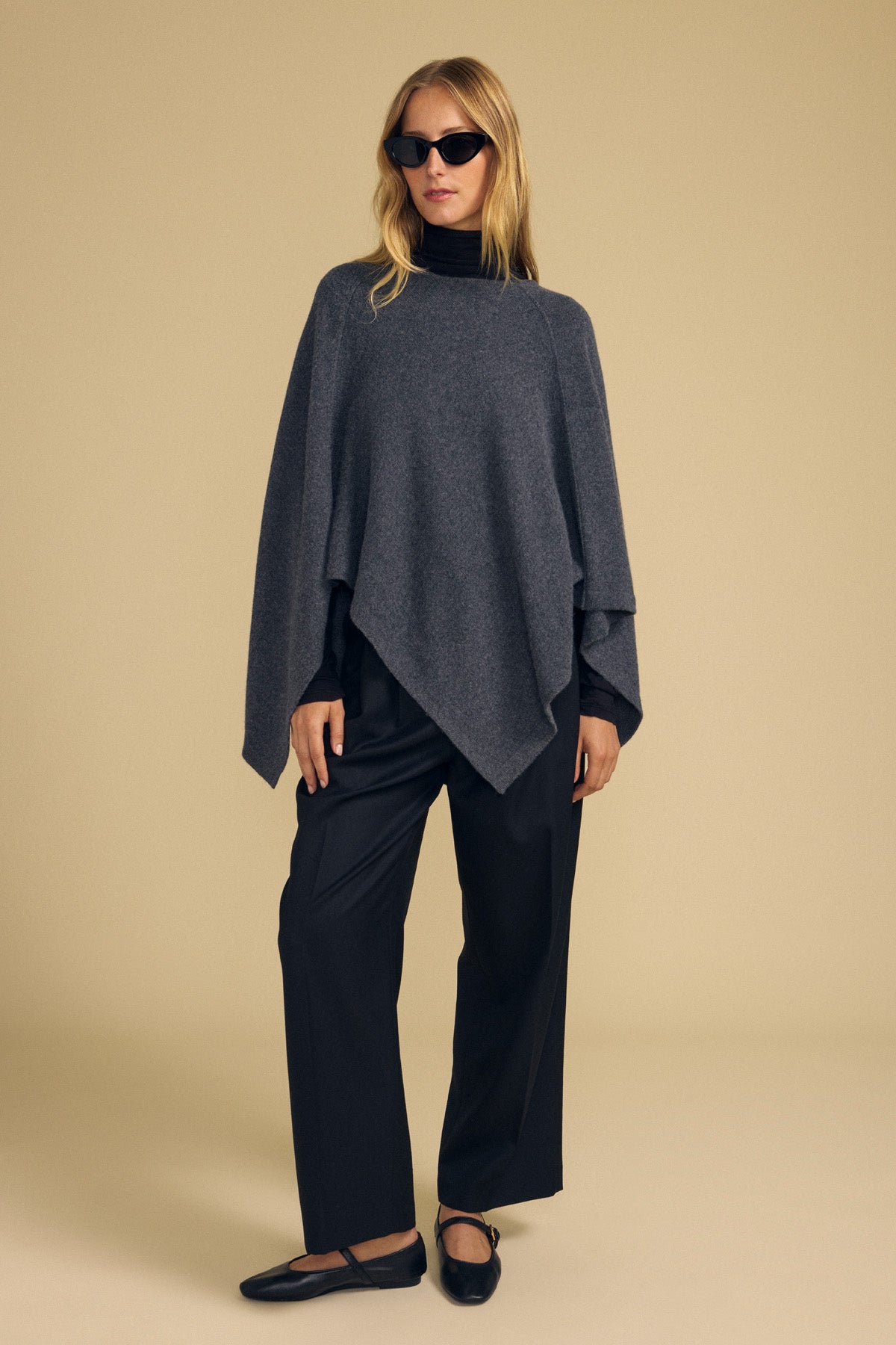 A person wearing the GIA PONCHO by Velvet by Graham & Spencer—a dark gray wool blend poncho—paired with a black turtleneck, loose black pants, black flats, and sunglasses stands against a plain beige background.-41960843018433