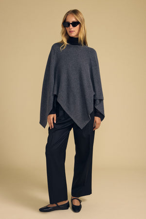 A person wearing the GIA PONCHO by Velvet by Graham & Spencer—a dark gray wool blend poncho—paired with a black turtleneck, loose black pants, black flats, and sunglasses stands against a plain beige background.