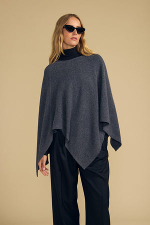 Wearing the GIA PONCHO by Velvet by Graham & Spencer—a dark gray wool blend style—paired with a black turtleneck, black wide-leg pants, and sunglasses, a person stands against a beige background.