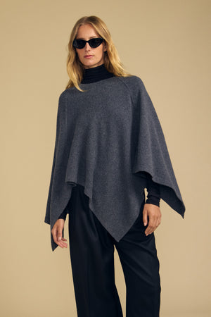 Draped in the GIA PONCHO by Velvet by Graham & Spencer, this person showcases effortless style with a soft gray merino wool layer over a black turtleneck and wide-leg pants. With long hair and sunglasses, they pose against a plain beige background. The versatile GIA PONCHO adds both comfort and sophistication to any look.