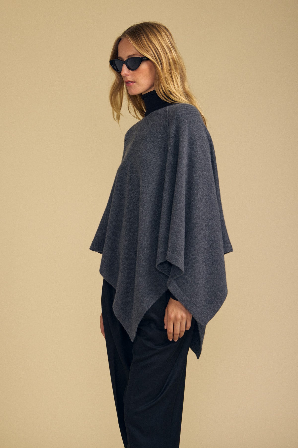 A person wearing black sunglasses, with the GIA PONCHO by Velvet by Graham & Spencer in dark gray layered over a black turtleneck and black pants, stands against a beige background.