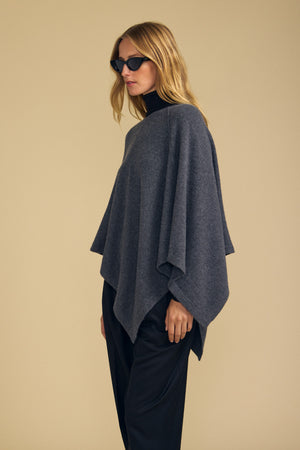 A person wearing black sunglasses, with the GIA PONCHO by Velvet by Graham & Spencer in dark gray layered over a black turtleneck and black pants, stands against a beige background.