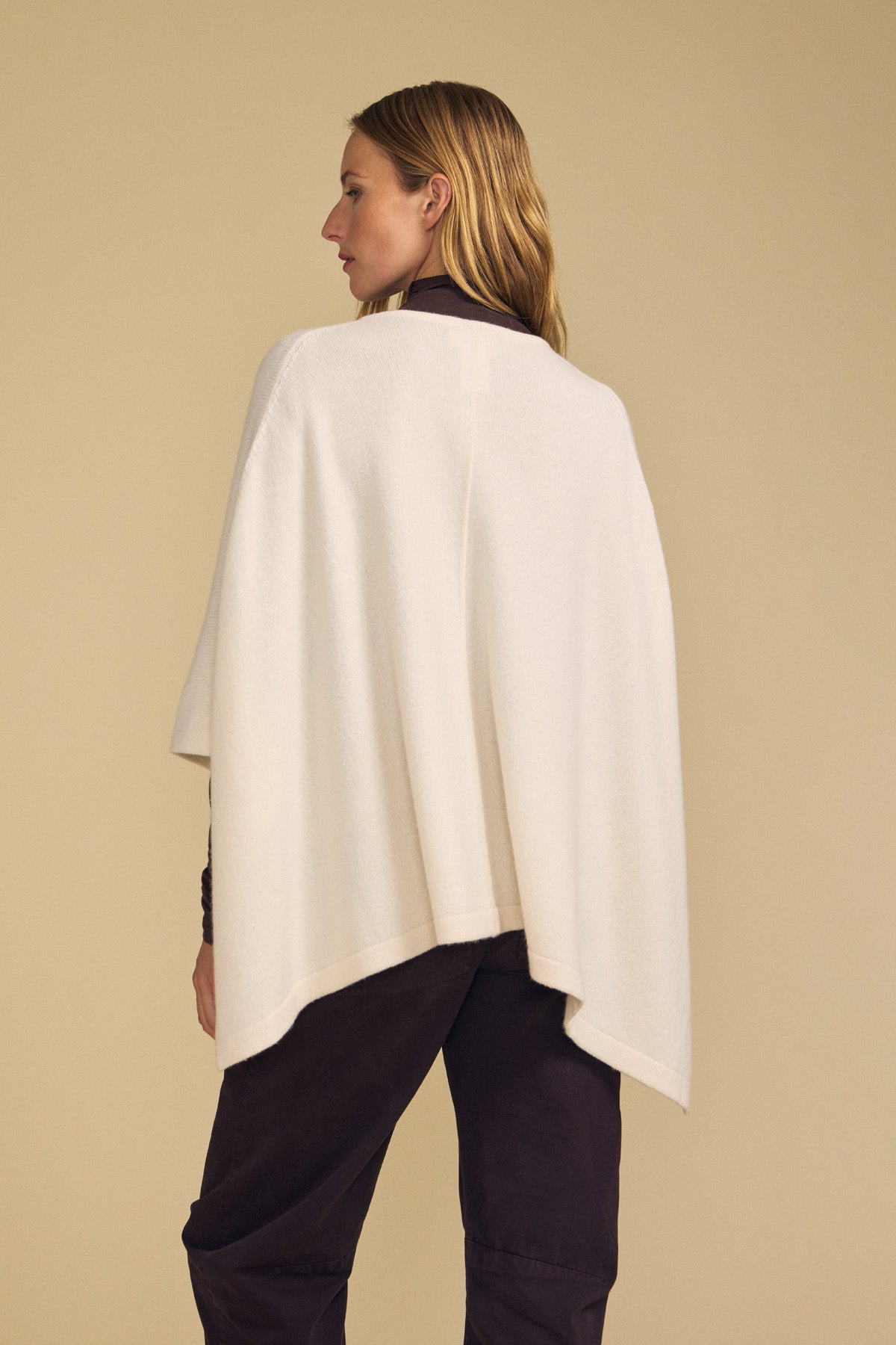 A person stands with their back to the camera, wearing the GIA PONCHO by Velvet by Graham & Spencer over a dark outfit, set against a beige background.
