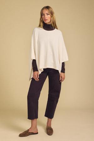A woman stands against a beige background wearing the GIA PONCHO from Velvet by Graham & Spencer, layered over a dark turtleneck and paired with cropped dark pants and brown loafers. This versatile piece adds both warmth and effortless style to her look.