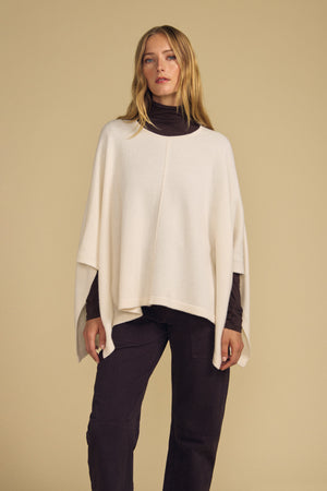 A woman stands against a beige background, wearing the GIA PONCHO in cream by Velvet by Graham & Spencer, layered over a dark turtleneck and dark pants.