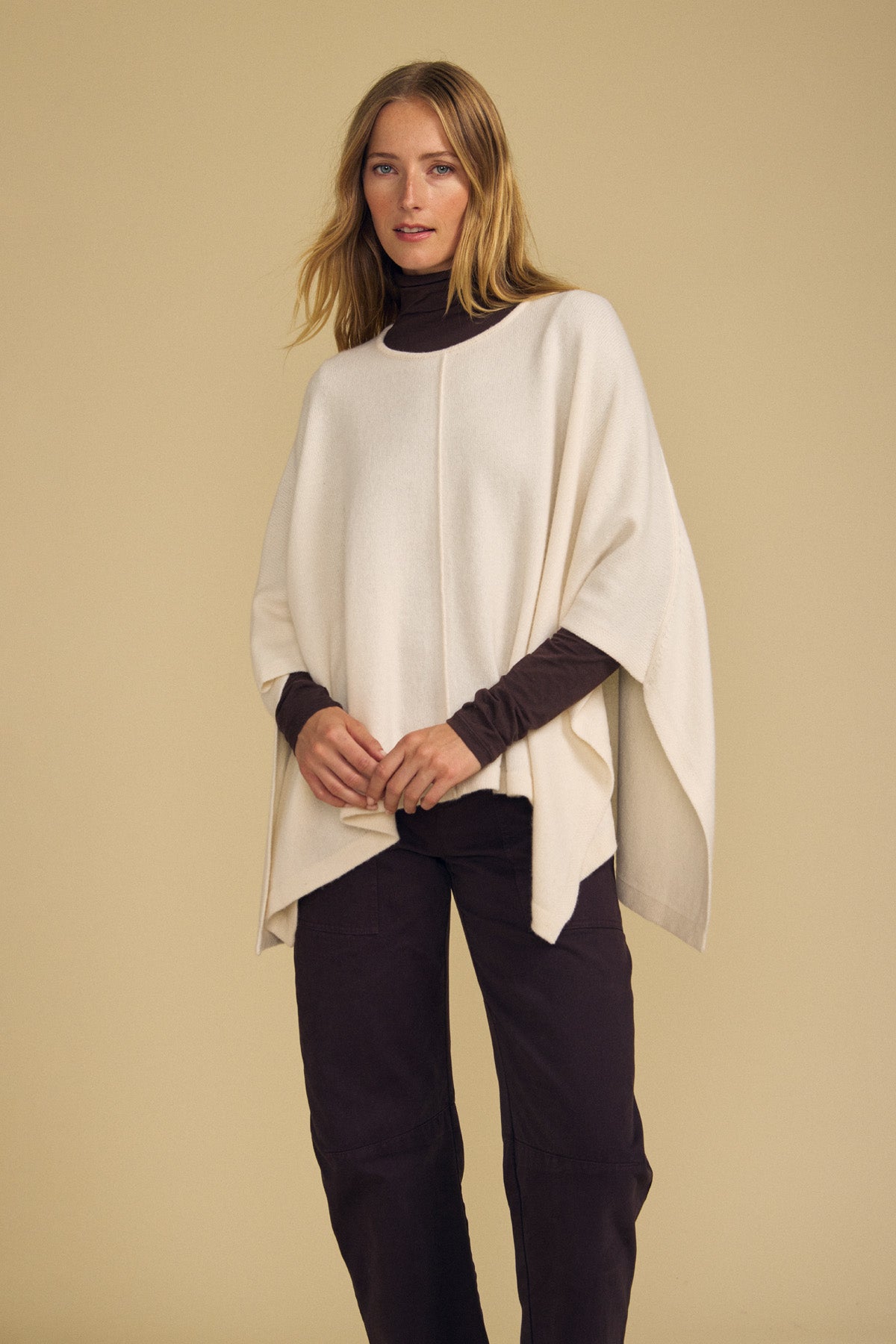 A woman wearing the GIA PONCHO by Velvet by Graham & Spencer in cream merino wool, layered over a brown turtleneck and brown pants, stands against a plain beige background.-41960843575489