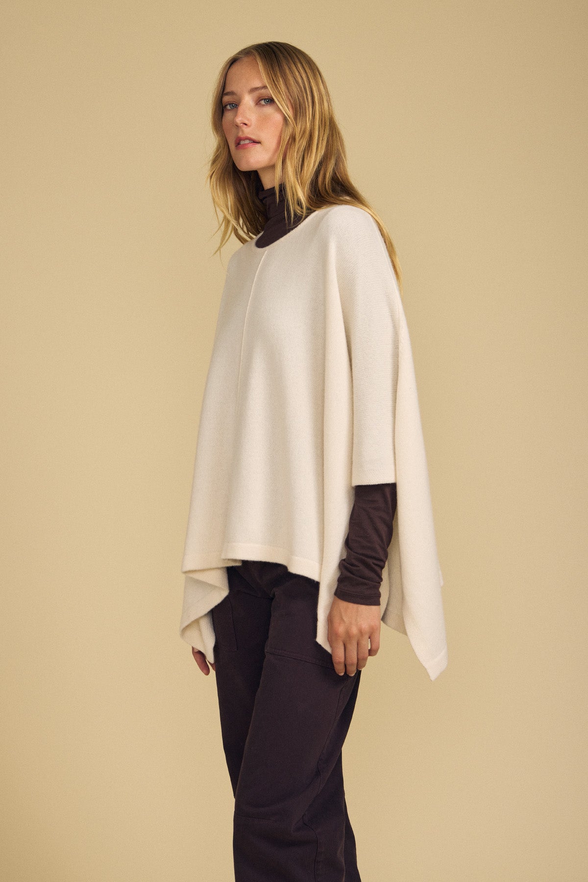 A woman stands against a beige background wearing the GIA PONCHO by Velvet by Graham & Spencer, layered over a dark long-sleeve shirt and dark pants—making it the perfect layering piece for cooler days.