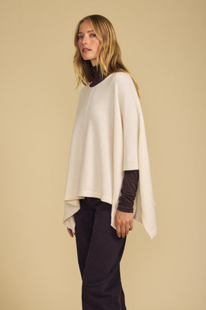 A woman stands against a beige background wearing the GIA PONCHO by Velvet by Graham & Spencer, layered over a dark long-sleeve shirt and dark pants—making it the perfect layering piece for cooler days.