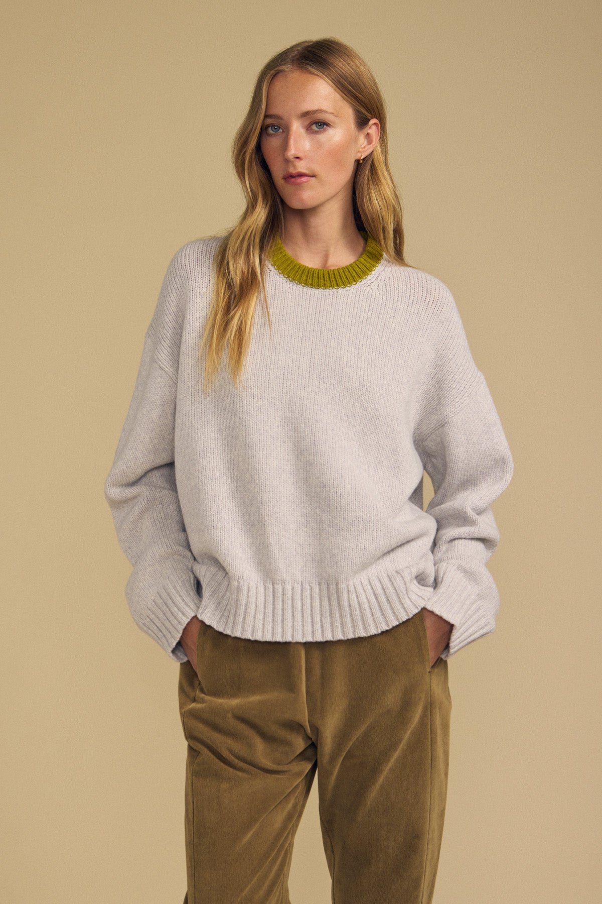 A woman stands with her hands in her pockets, wearing the Velvet by Graham & Spencer CALEN SWEATER—a light gray crewneck sweater featuring a green neckline—paired with olive pants, set against a beige background.