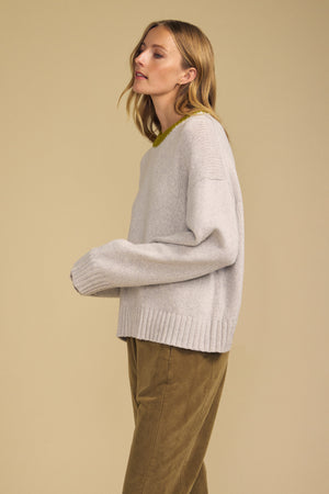 A person stands sideways against a beige background wearing the CALEN SWEATER by Velvet by Graham & Spencer—a cozy light gray women’s crewneck with ribbed cuffs and hem—paired with olive green pants.