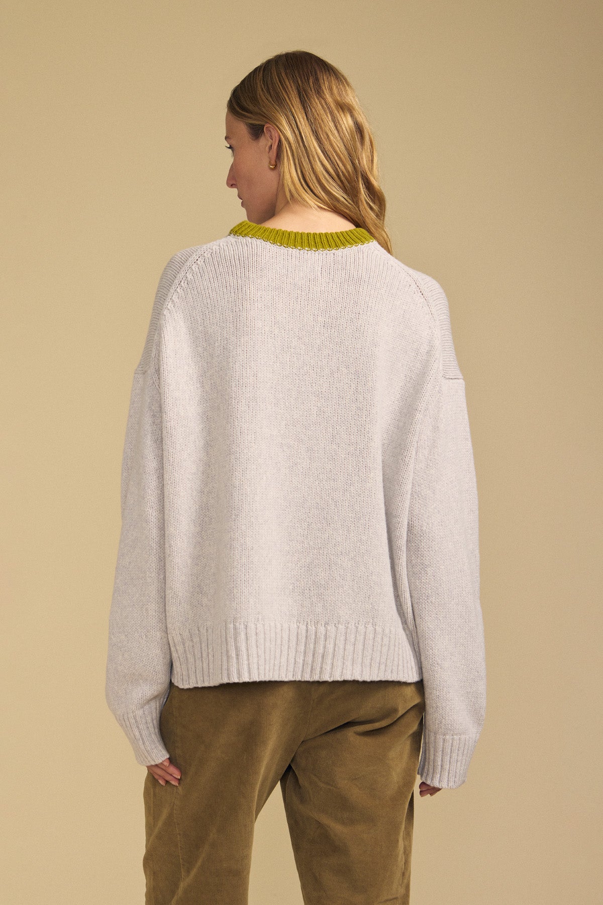 A person stands with their back to the camera, wearing the CALEN SWEATER by Velvet by Graham & Spencer—a light gray women’s crewneck sweater featuring a yellow collar—paired with tan pants, set against a plain beige background.