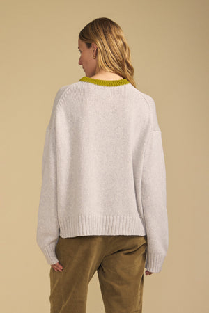 A person stands with their back to the camera, wearing the CALEN SWEATER by Velvet by Graham & Spencer—a light gray women’s crewneck sweater featuring a yellow collar—paired with tan pants, set against a plain beige background.