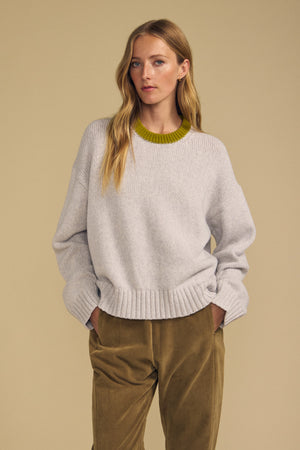 A woman stands with her hands in her pockets, wearing the Velvet by Graham & Spencer CALEN SWEATER—a light gray crewneck sweater featuring a green neckline—paired with olive pants, set against a beige background.