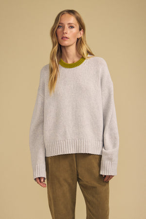 A woman stands against a beige background, wearing the CALEN SWEATER by Velvet by Graham & Spencer—a women’s crew neck sweater in light gray with a green collar detail—paired with olive trousers. This cozy CALEN SWEATER, crafted from soft material, blends effortless style and comfort for an elevated everyday look.