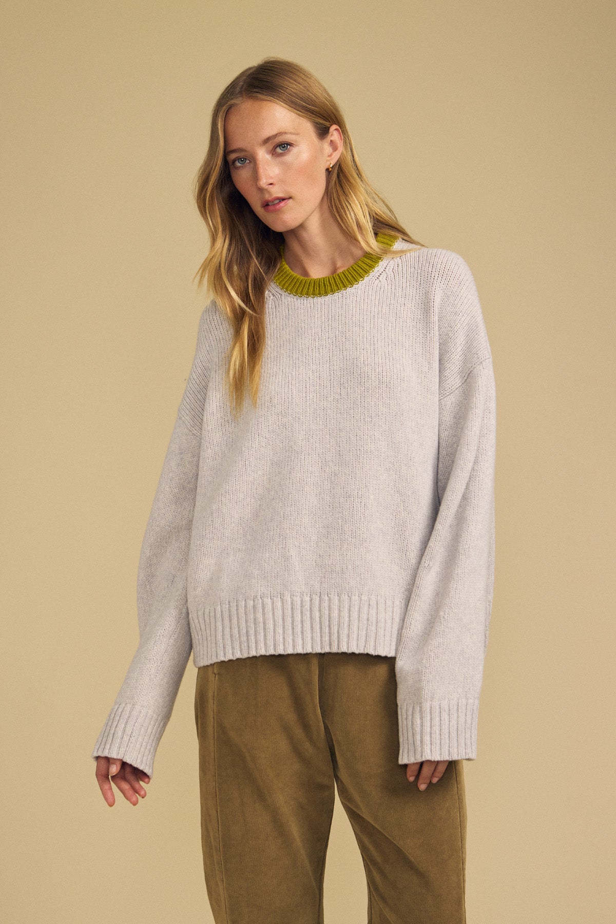 A woman wearing the CALEN SWEATER by Velvet by Graham & Spencer—a cozy women’s crewneck in light gray with a green neckline—paired with tan pants, stands against a plain beige background.-42063732441281