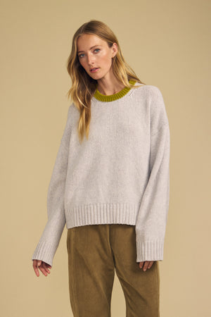 A woman wearing the CALEN SWEATER by Velvet by Graham & Spencer—a cozy women’s crewneck in light gray with a green neckline—paired with tan pants, stands against a plain beige background.