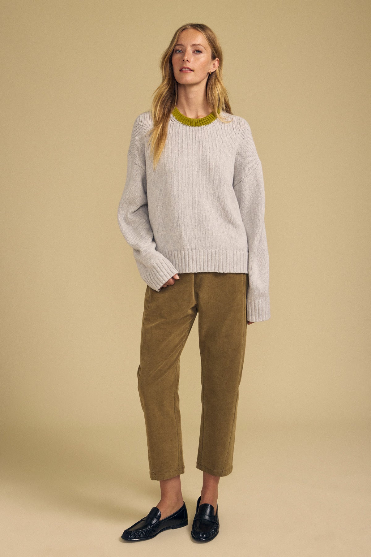 Wearing the CALEN SWEATER by Velvet by Graham & Spencer in cozy light gray, paired with olive corduroy pants and black loafers, a woman stands against a beige background looking forward with a relaxed posture.