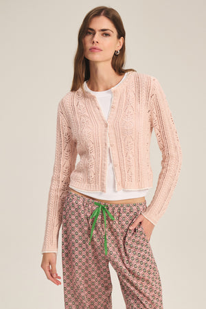 Woman wears the DAISY CARDIGAN by Velvet by Graham & Spencer, a light pink cotton-blend piece with pointelle details, layered over a white top and patterned drawstring pants with a green tie, against a plain background.
