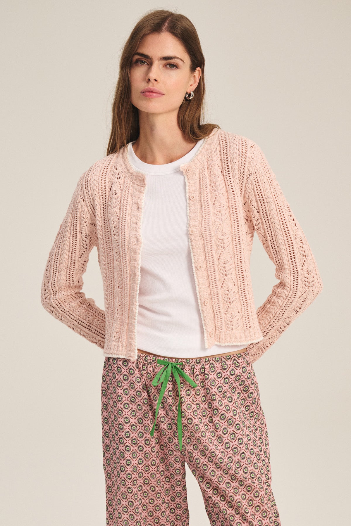 A woman wears the DAISY CARDIGAN by Velvet by Graham & Spencer in light pink over a white t-shirt and patterned pants with a green drawstring, standing against a plain background.-41460002095297