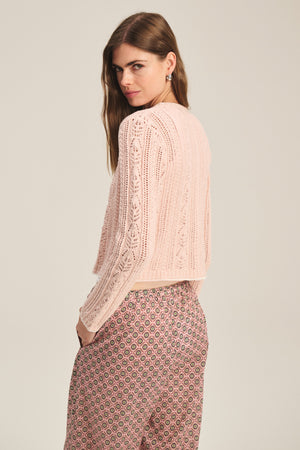 A woman with long brown hair wears the DAISY CARDIGAN by Velvet by Graham & Spencer in light pink cotton blend, paired with patterned pink pants, standing sideways and looking over her shoulder.