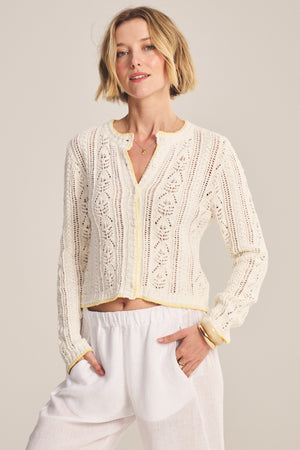 A woman wears the DAISY CARDIGAN by Velvet by Graham & Spencer—a cream, cropped knit with pointelle and scalloped edges—paired with loose white pants, standing against a plain background.