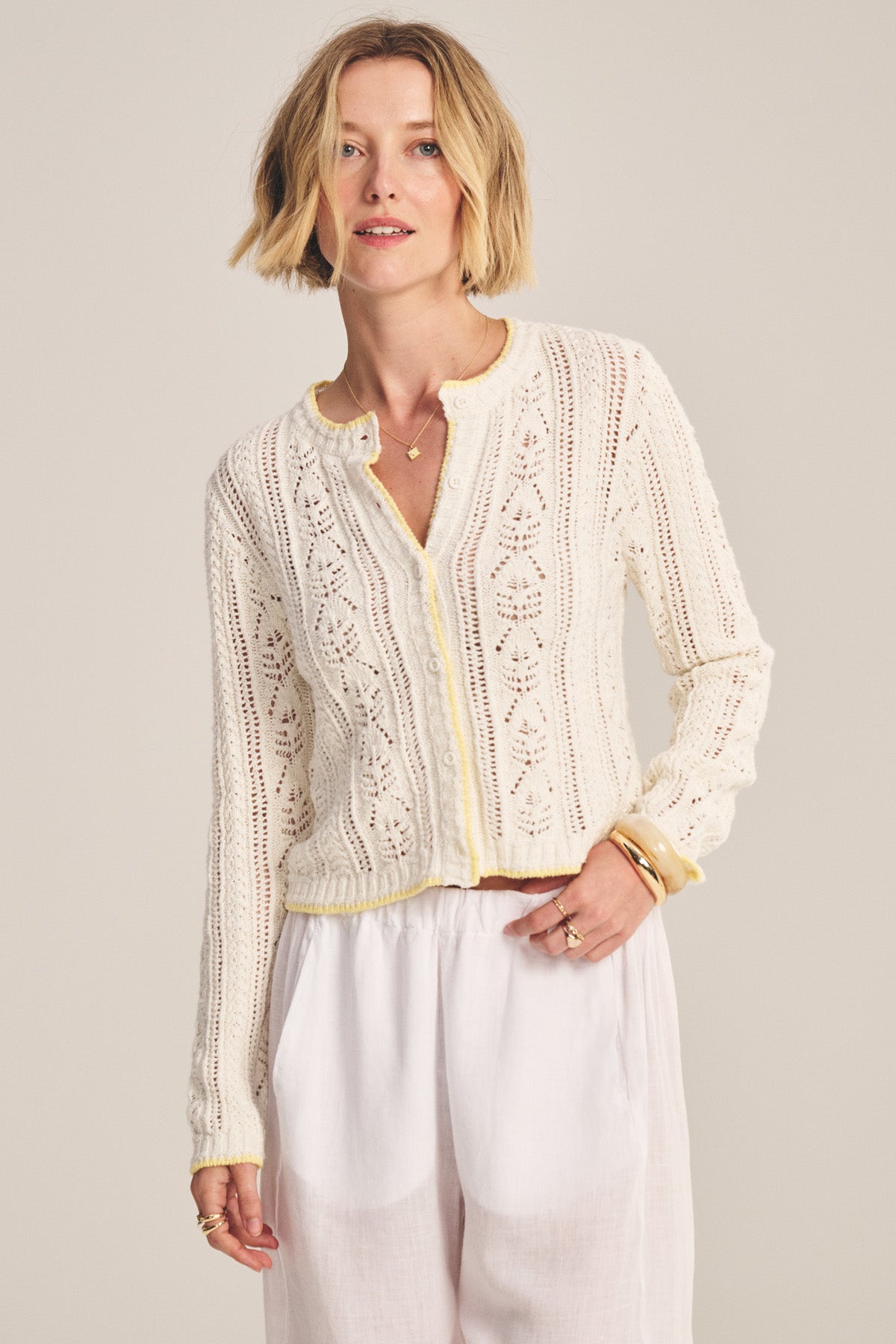 A woman with short blonde hair models the DAISY CARDIGAN by Velvet by Graham & Spencer, a white knit cropped cardigan with yellow trim and subtle pointelle details, styled with white pants against a neutral background.