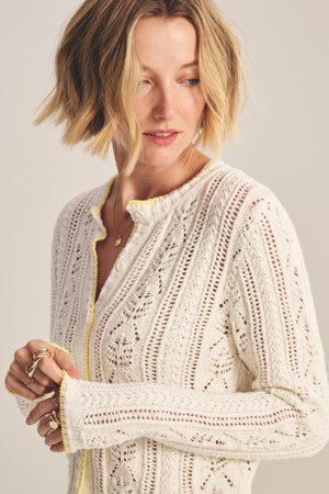 A woman with short blonde hair models the DAISY CARDIGAN by Velvet by Graham & Spencer, a white cropped style with subtle yellow trim, paired with gold jewelry as she looks slightly to her left.