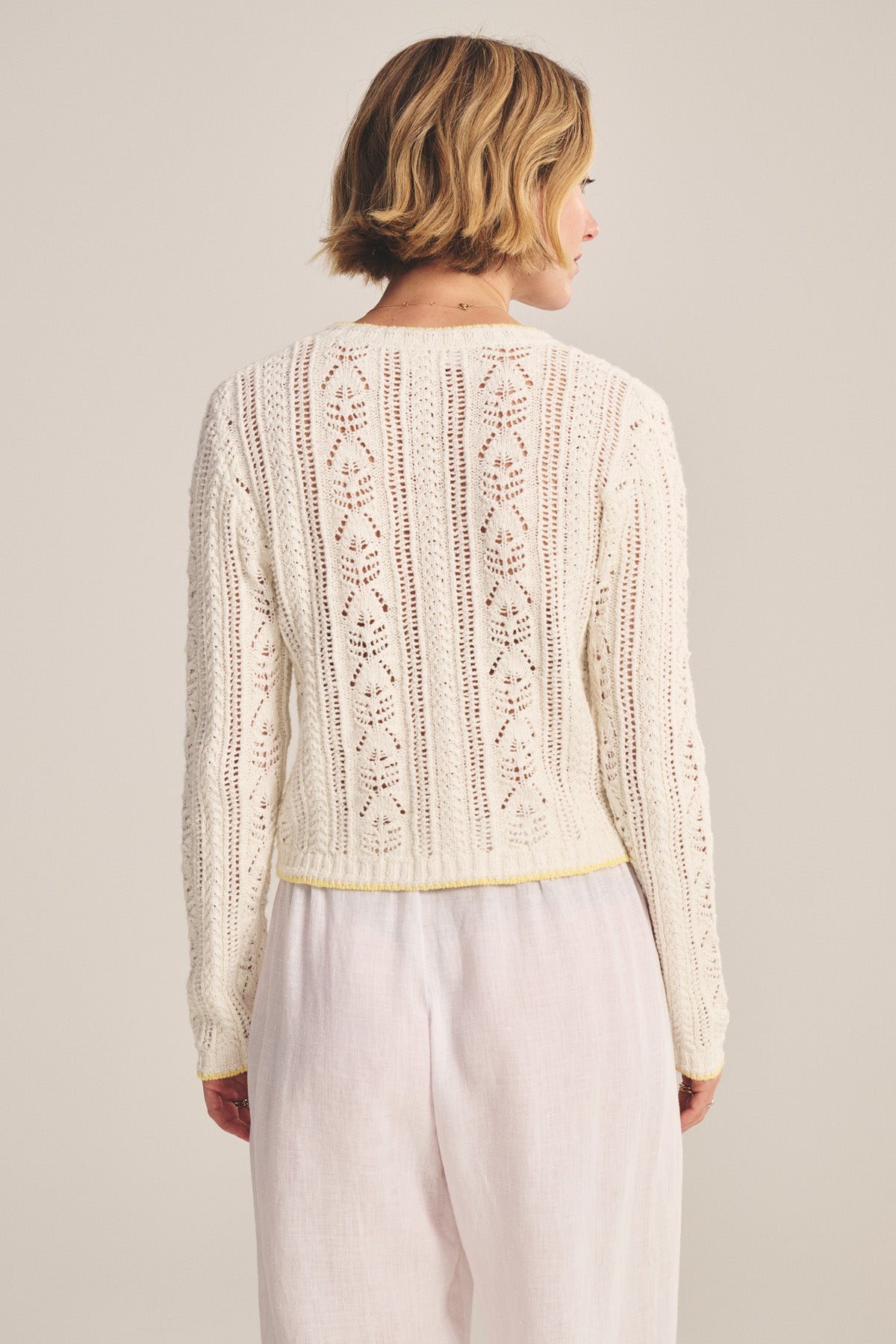 A person with short blonde hair is seen from the back, wearing Velvet by Graham & Spencer's DAISY CARDIGAN, a long-sleeved white knit with pointelle detailing, paired with white pants.
